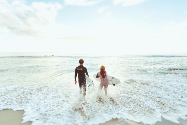 Two people are walking into the ocean while holding surfboards, with waves crashing onto the shore and a partly cloudy sky in the background.