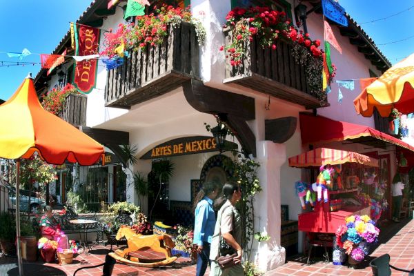 This image shows a vibrant marketplace with colorful umbrellas, banners, flowers, and a couple walking by a store named "Artes de Mexico."