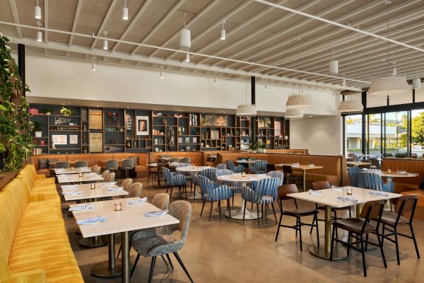 The image shows a modern, spacious restaurant interior with tables, chairs, cushions, and a wall with shelves.