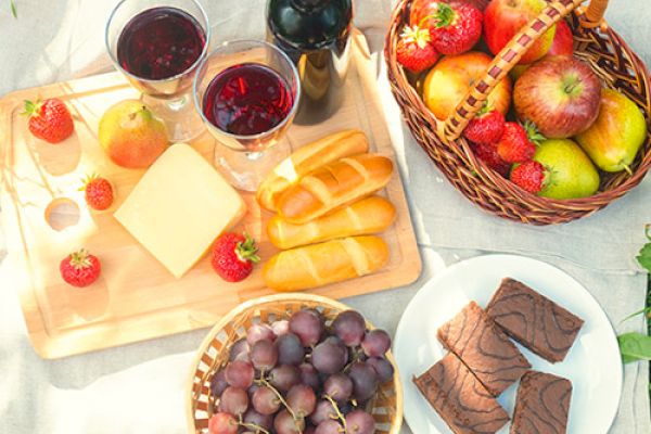 The image shows a picnic setup with wine, bread, cheese, grapes, strawberries, apples, pears, and chocolate brownies on a blanket.