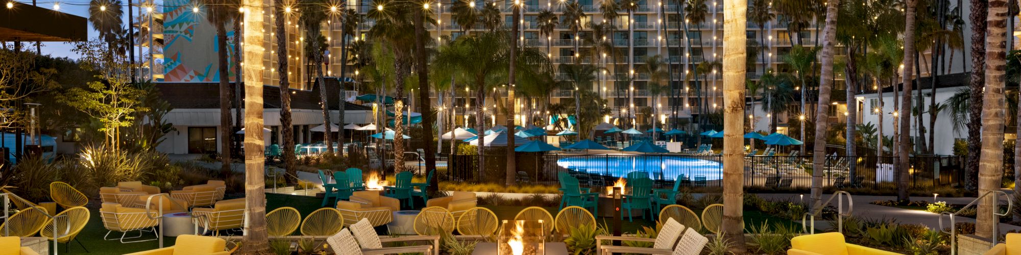 The image depicts an outdoor lounge area with fire pits, seating, and string lights, set against a backdrop of palm trees and a poolside resort.