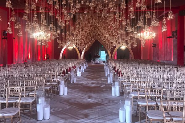 An ornate indoor venue set up for an event with rows of white chairs, hanging chandeliers, and glowing arches, creating an elegant and festive ambiance.