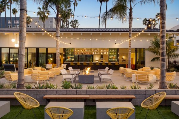 This image shows an outdoor patio with modern seating, string lights, palm trees, and a central fire pit, all facing a bar with stools.