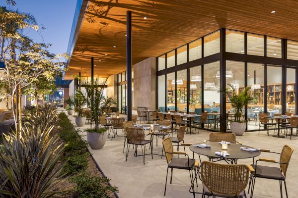 An outdoor dining area with tables and chairs, surrounded by potted plants, located next to a modern building with large glass windows, illuminated at dusk.