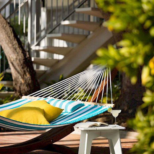 The image shows a colorful hammock with pillows, a small white table, and greenery, set in an outdoor area with a staircase in the background.