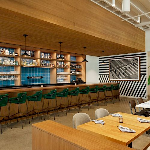 The image shows a modern bar with green barstools, a wooden counter, shelves stocked with bottles, and a small dining area with tables and chairs.
