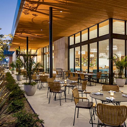 An outdoor restaurant with modern decor, potted plants, and wicker chairs. The space is well-lit and inviting, with large windows and an airy ambiance.