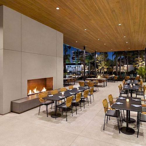 An outdoor dining area with modern furniture and a lit fireplace, featuring tables and chairs set for guests, under a wooden ceiling at night.