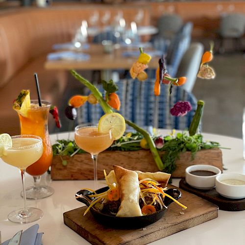 The image shows an elegant table setting with cocktails, a vibrant salad with colorful skewered items, and a dish served on a wooden board with side sauces.