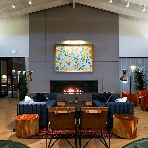 A modern living room with a central fireplace, abstract art above, blue couches, orange chairs, wooden furniture, and a high ceiling with exposed beams.
