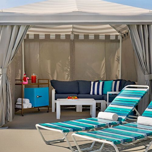 Outdoor cabana with a couch, pillows, table, and two striped lounge chairs. A side table holds a pink flamingo and a drink.