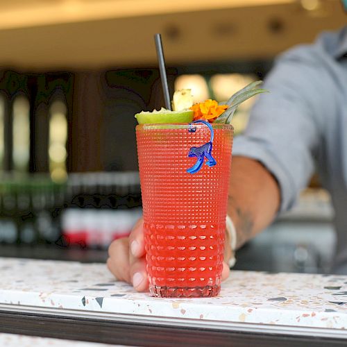 A person wearing a mask holds a red beverage adorned with tropical garnishes, including a pineapple slice and a blue stirrer, at a bar.