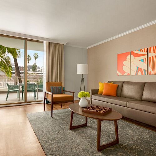 This image shows a modern living room with a sofa, coffee table, flat-screen TV, and a vibrant painting. There's a balcony with a table and chairs.
