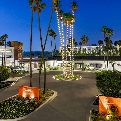 A well-lit, scenic outdoor area with tall palm trees, a roundabout, modern buildings, and a mural of a colorful sun on a wall at dusk or nighttime.