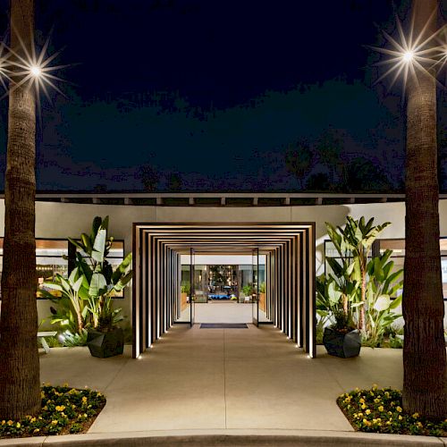 This image shows a modern entrance with illuminated pathways, flanked by palm trees and lush greenery at night, leading into a well-lit building.