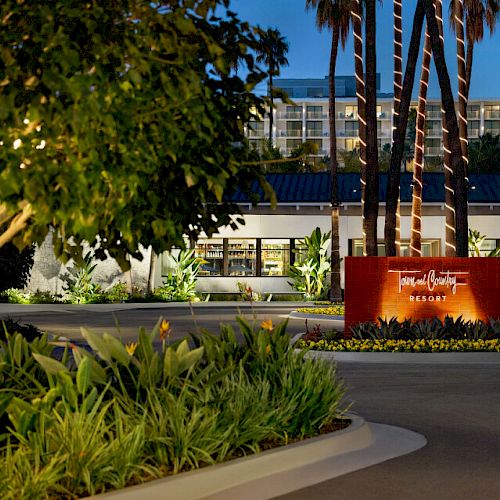 The image shows an entrance to a resort at dusk with a sign, surrounded by palm trees and lush greenery. The scene is well-lit with ambient lighting.