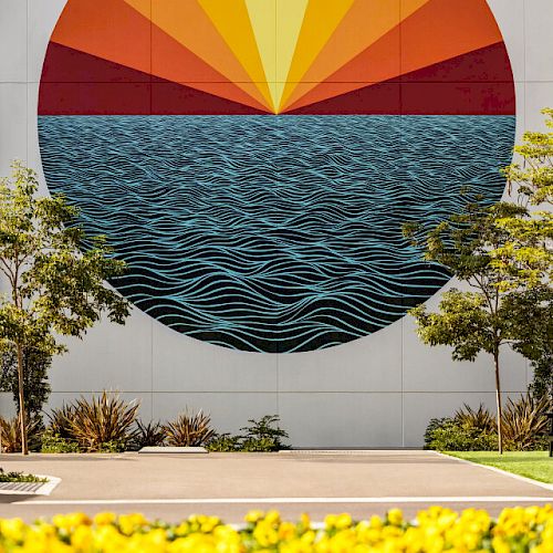 The image shows a mural of a stylized sunset over the ocean, with yellow flowers and trees in the foreground, painted on a building wall.