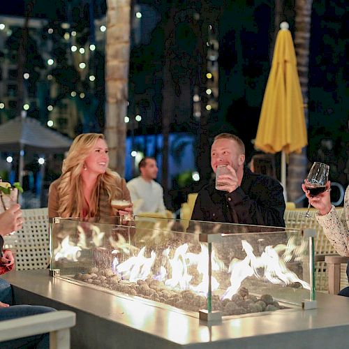 A group of people sits around a fire pit outdoors at night, enjoying drinks and smiling, with lights and trees in the background.