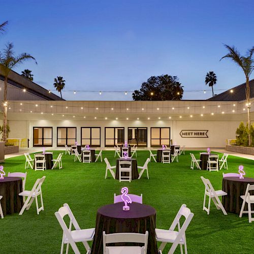 An outdoor venue with round tables, white chairs, and string lights set up on a green lawn, surrounded by buildings and palm trees.