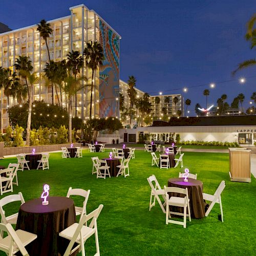 An outdoor event space is set up with round tables and chairs on a grassy area, decorated with purple centerpieces, under string lights and palm trees.