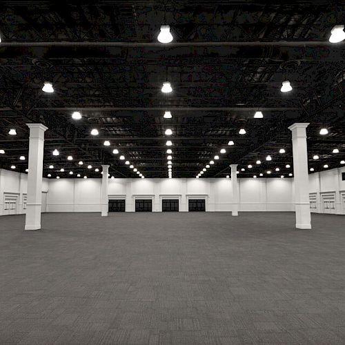 This image shows an empty, large, well-lit exhibition hall with a high ceiling, numerous overhead lights, columns, and a gray carpeted floor.