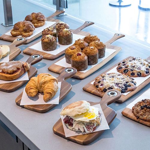 The image shows a variety of pastries, muffins, cookies, croissants, and sandwiches displayed on wooden boards atop a counter.