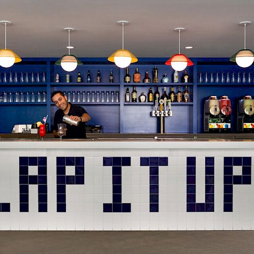 A bartender prepares a drink behind a modern bar with "LAP IT UP" written in blue tiles on the counter.