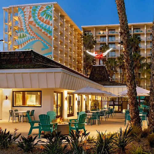 The image shows an outdoor seating area with teal chairs and tables, palm trees, a building with a colorful mural, and another taller hotel building.