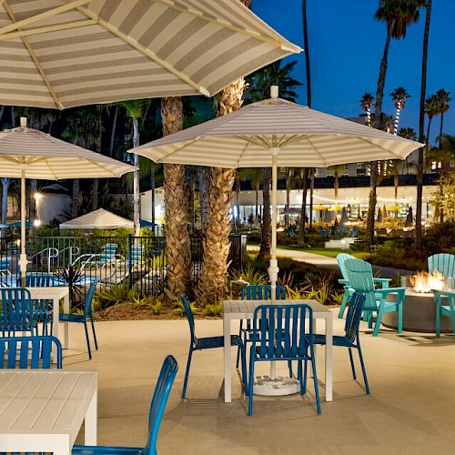 The image shows an outdoor patio area with tables, chairs, and umbrellas, near a pool, surrounded by palm trees and evening lighting.