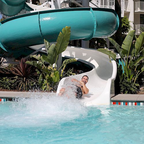 The image shows a person sliding down a water slide into a pool, surrounded by green plants and some buildings in the background.