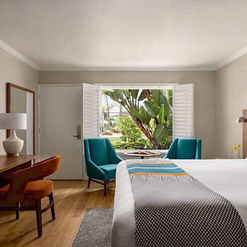 A hotel room featuring a bed, two chairs by the window, a desk with a chair, a dresser with a TV, artwork, and a garden view through the window.