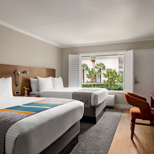 A hotel room with two double beds, a window with a view of palm trees, a dresser with a TV and lamp, and a chair.