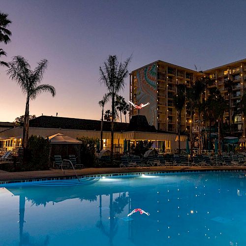 A large outdoor swimming pool at dusk, surrounded by lounge chairs and palm trees, with a multi-story hotel in the background.