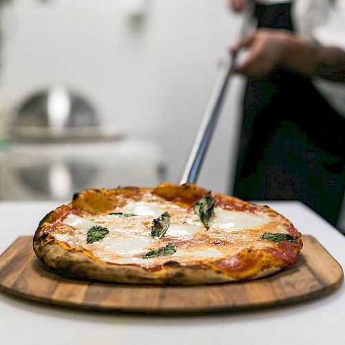 A fresh pizza topped with melted cheese and basil on a wooden board, being served by a person in a kitchen.