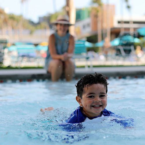 A smiling child is swimming in a pool, with an adult sitting poolside in the background on a sunny day with some chairs and umbrellas visible.