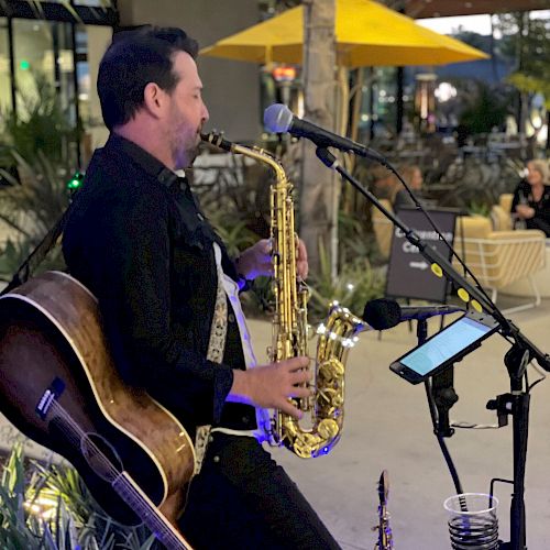 A musician playing the saxophone, with a guitar strapped to his back, performing at an outdoor venue under string lights and a yellow umbrella.