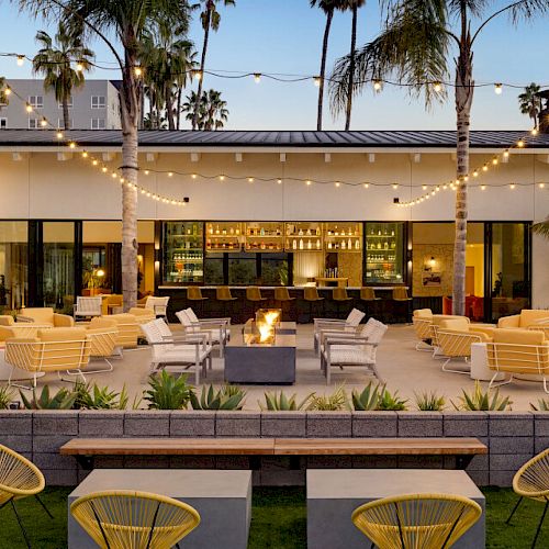 The image features an outdoor patio with comfortable seating, string lights, a fire pit, and a well-lit bar area, surrounded by palm trees and modern decor.
