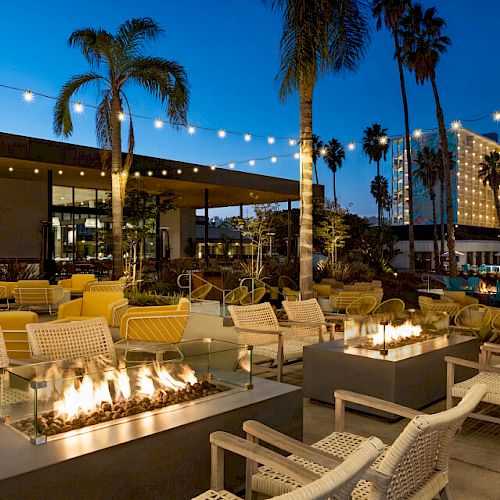 An outdoor lounge area with fire pits, cushioned seating, string lights, palm trees, and surrounding buildings illuminated against a twilight sky.