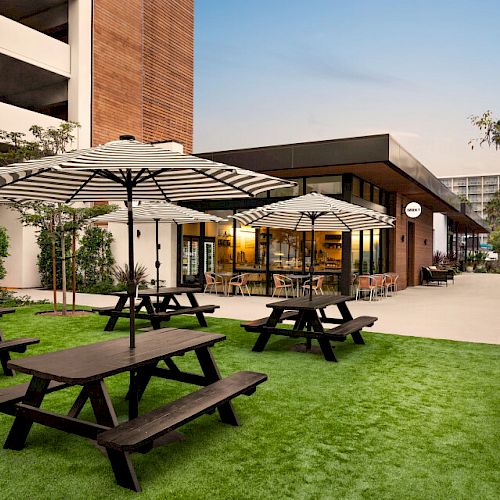 An outdoor seating area with picnic tables and striped umbrellas on a grass surface adjacent to a modern building with large windows.