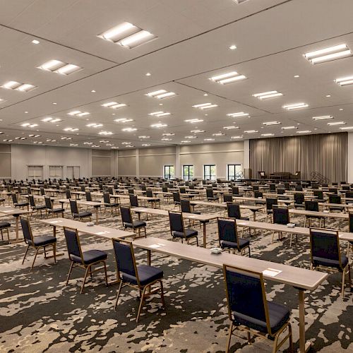 A large, well-lit conference room with rows of tables and chairs arranged neatly, ready for an event or meeting. The room has a contemporary design.