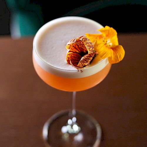 A cocktail in a coupe glass, featuring a frothy top, garnished with a slice of dried citrus and a yellow edible flower, placed on a wooden surface.
