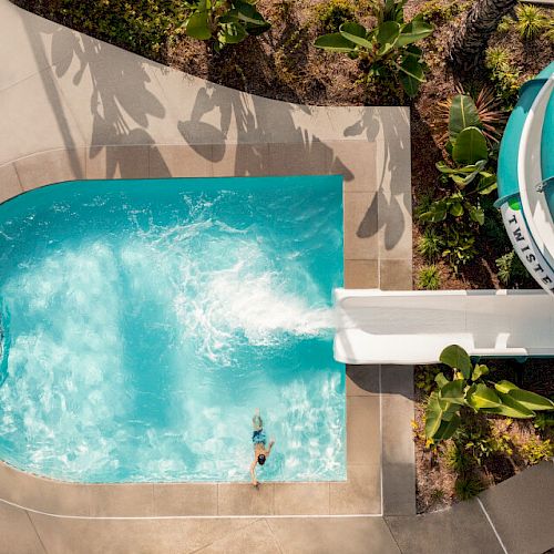 Aerial view of a water slide leading into a small pool, with one person swimming, surrounded by greenery and concrete pathways, during daylight.