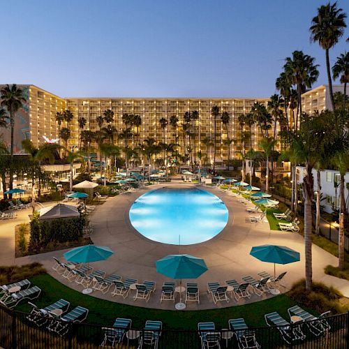 The image shows a luxurious resort with a large circular pool, surrounded by lounge chairs and palm trees, and a hotel building in the background.