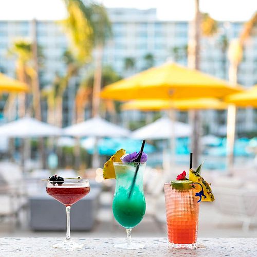Three colorful cocktails sit on a marble table with a blurred background of yellow umbrellas and palm trees in a tropical setting.