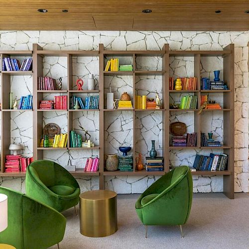 A colorful, cozy reading area features green chairs, a round table, a bookshelf with colorful items, and a lamp in a bright, well-lit room.