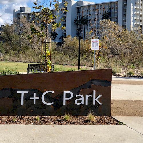 A sign for T+C Park is in the foreground with a modern building and some trees in the background. The weather appears to be partly cloudy.