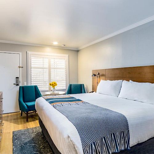 A modern hotel room featuring a large bed with white linens, two blue chairs, a wooden dresser, and a flat-screen TV in front of the bed.