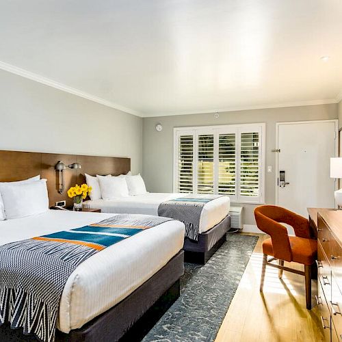 A modern hotel room with two queen beds, a white desk and chair, a flatscreen TV on a wooden dresser, yellow flowers, and a window in the background.