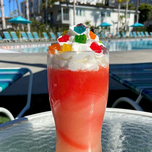 A colorful drink topped with whipped cream and gummy bears is on a table by a pool. Palm trees and buildings can be seen in the background.