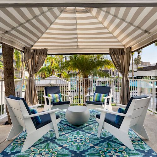 A shaded outdoor seating area with four white chairs, a round table, and a blue-patterned rug, surrounded by a fence and palm trees.